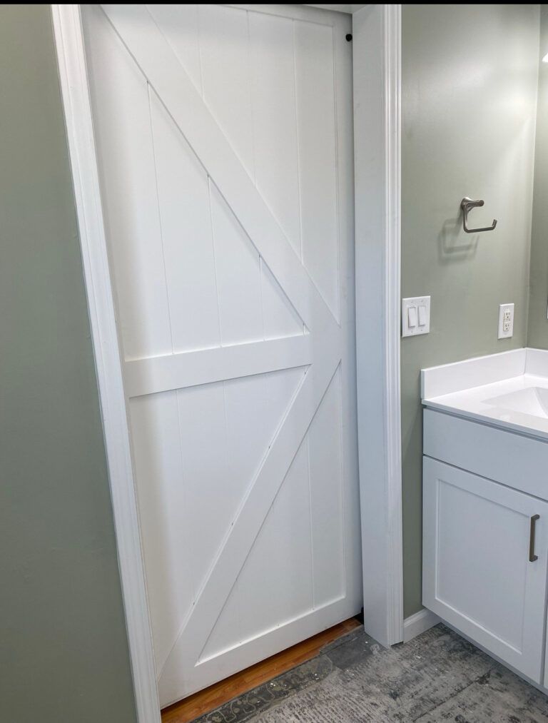 White farmhouse-style sliding door partially open to a bathroom. The bathroom has a white vanity and light green walls.