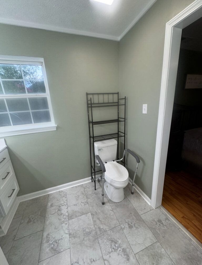 Bathroom with toilet and assistive grab bars. A shelf unit is over the toilet, window to the left, and doorway on the right.
