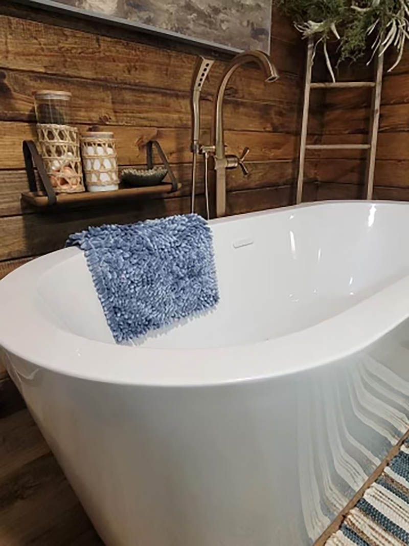 A white freestanding bathtub with a blue bath mat inside, set in a rustic bathroom with wood paneling.