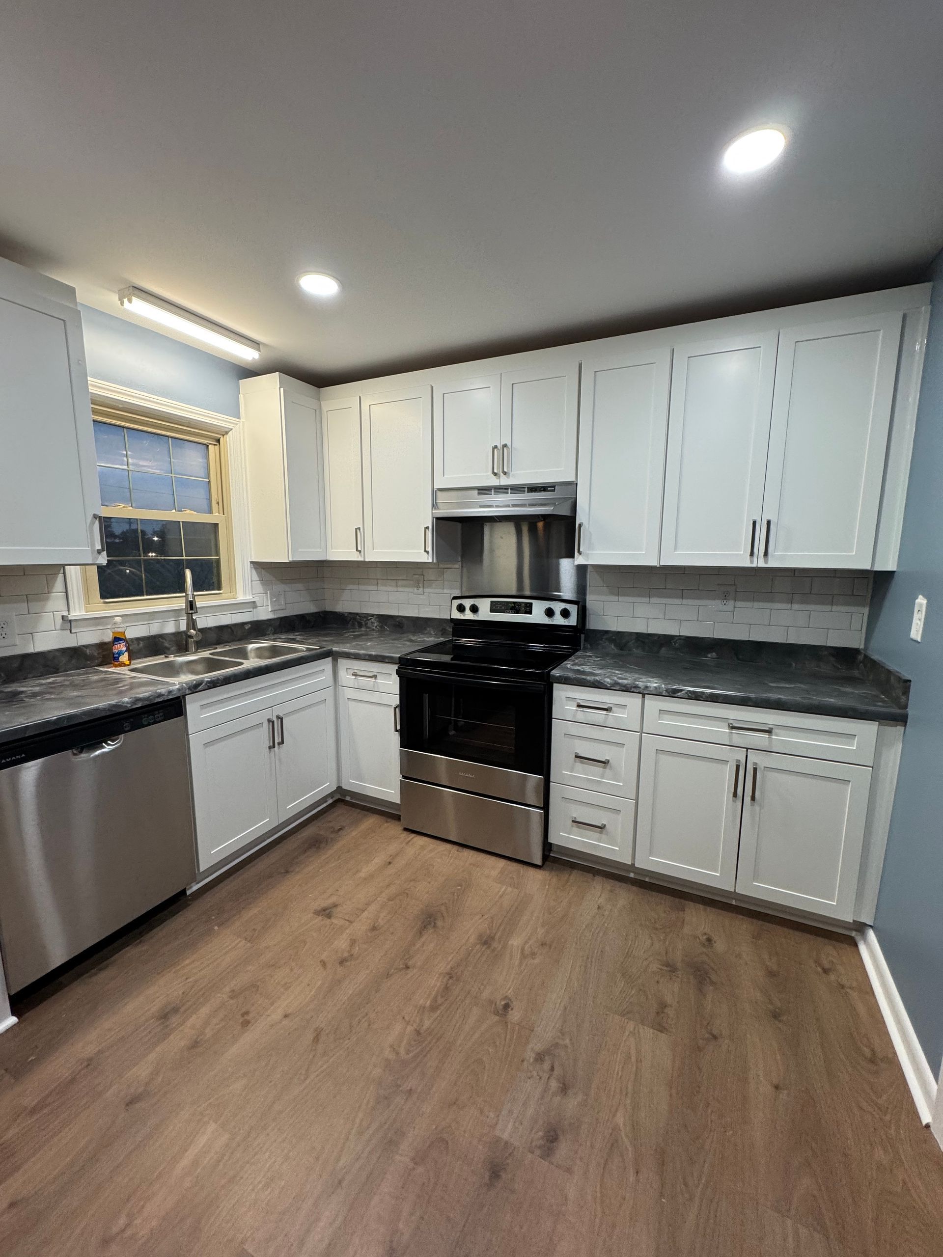 A renovated kitchen with white cabinets, stainless steel appliances, dark countertops, and wood-look flooring.