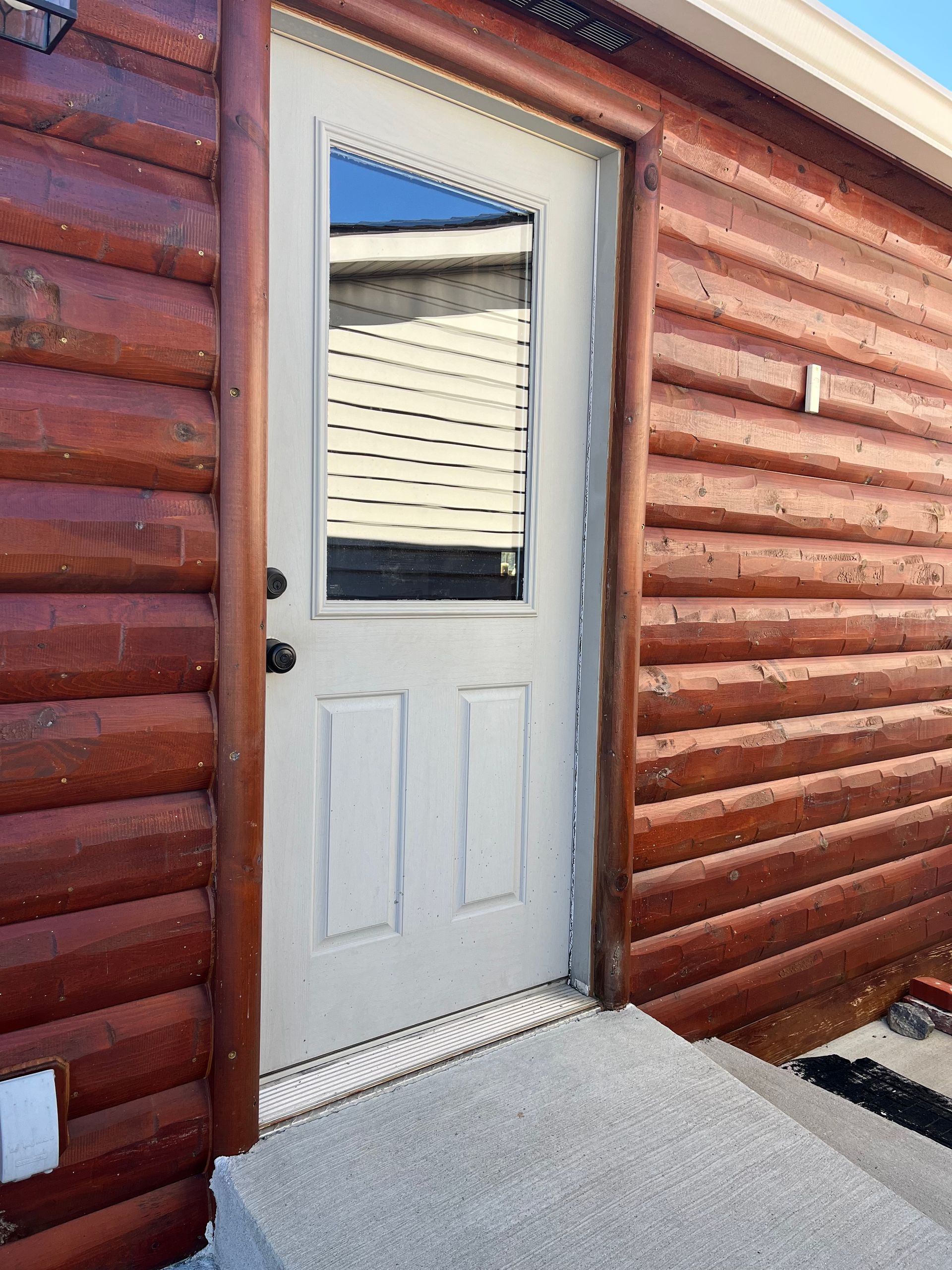 White door with glass pane in red log cabin wall; concrete steps lead to entrance.