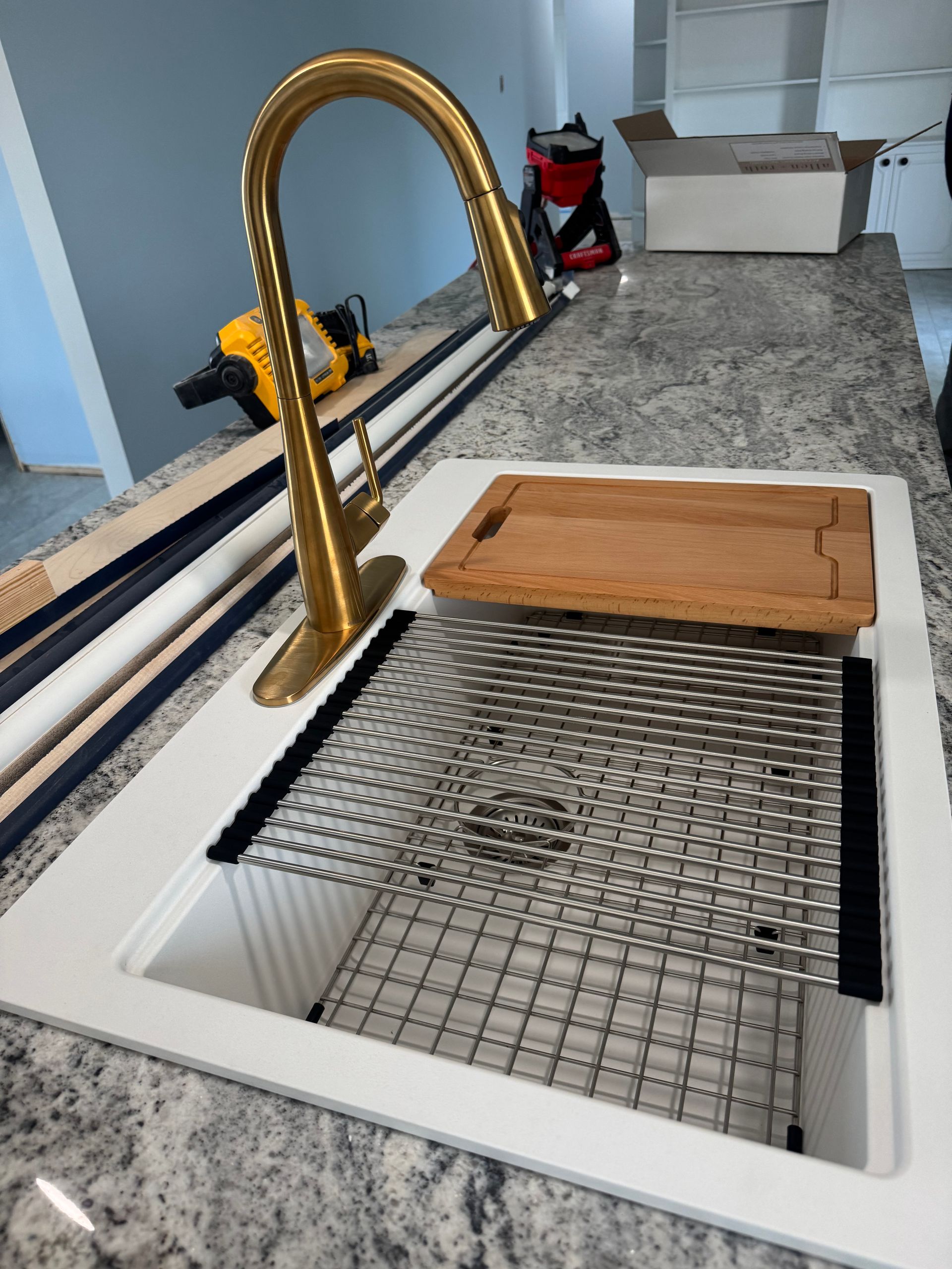 Gold faucet over a white sink with a metal grate and wooden cutting board, on a granite countertop.