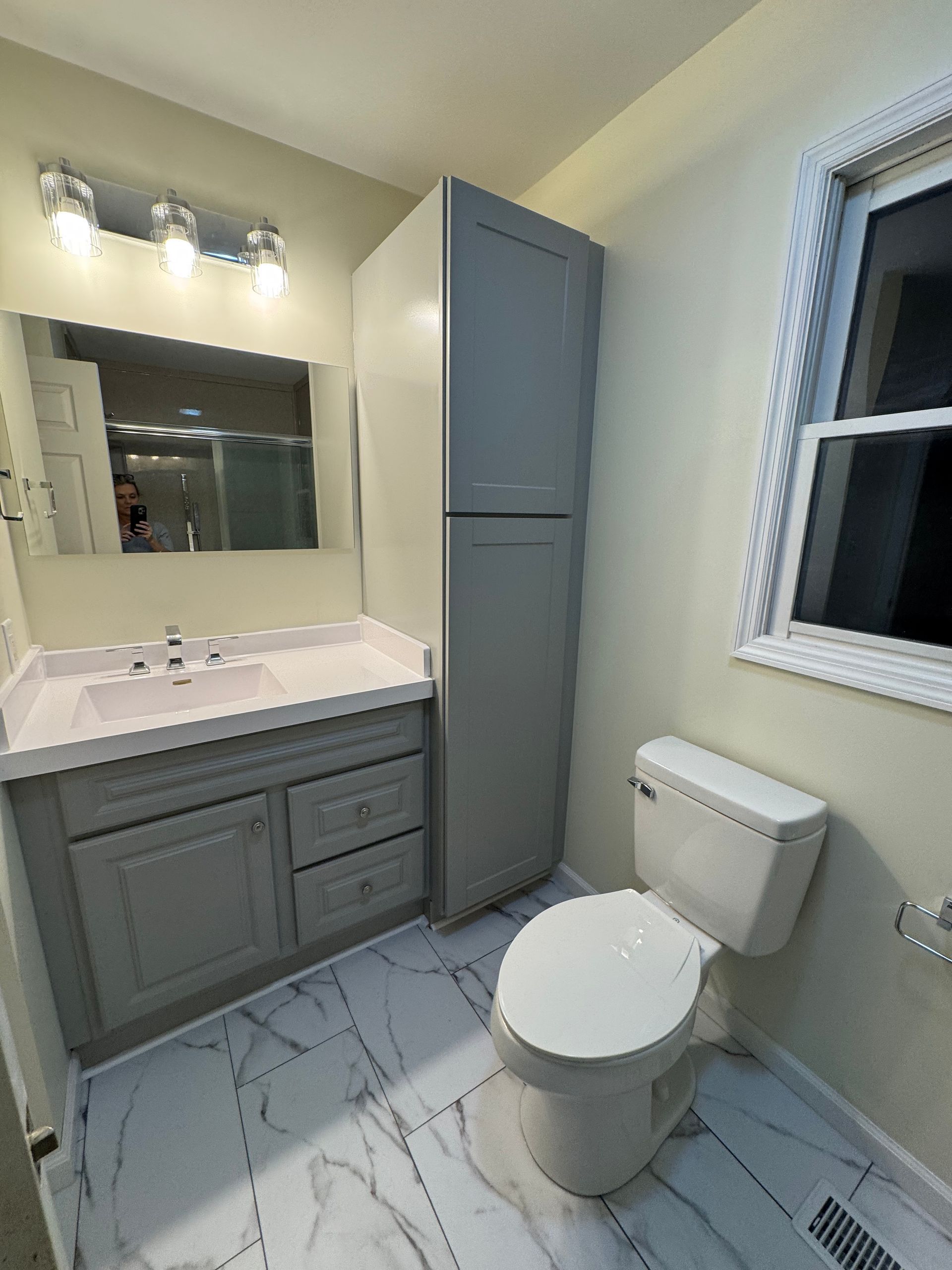 Bathroom with gray cabinetry, white toilet, marble-look flooring, and a window.