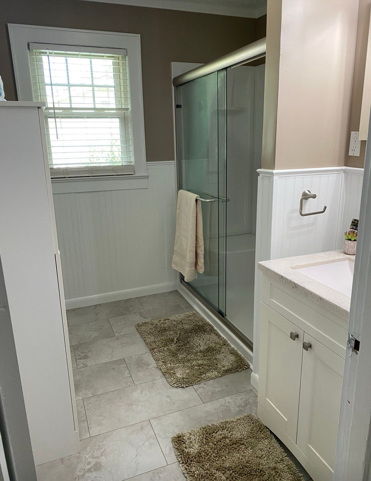 Bathroom with a shower, vanity, window, and two rugs. Gray tile floor, white cabinets and wainscoting, tan walls.