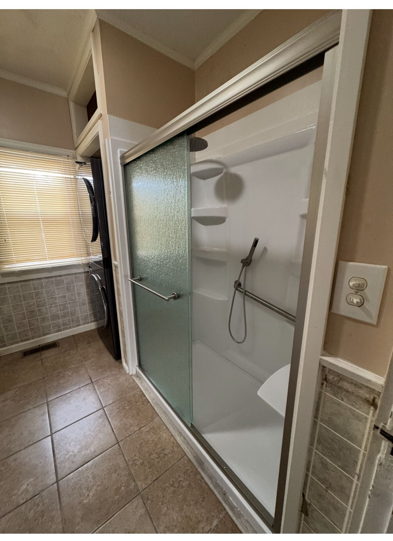 Bathroom with a white-framed shower, glass door, grab bar, and built-in seat. Brown tile floor, tan walls.