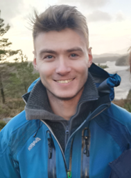 Mann iført blå jakke, sjø og fjell, kystlandskap i bakgrunnen. Foto.