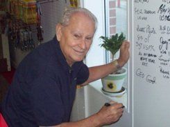 An elderly man is writing on a whiteboard next to a potted plant.