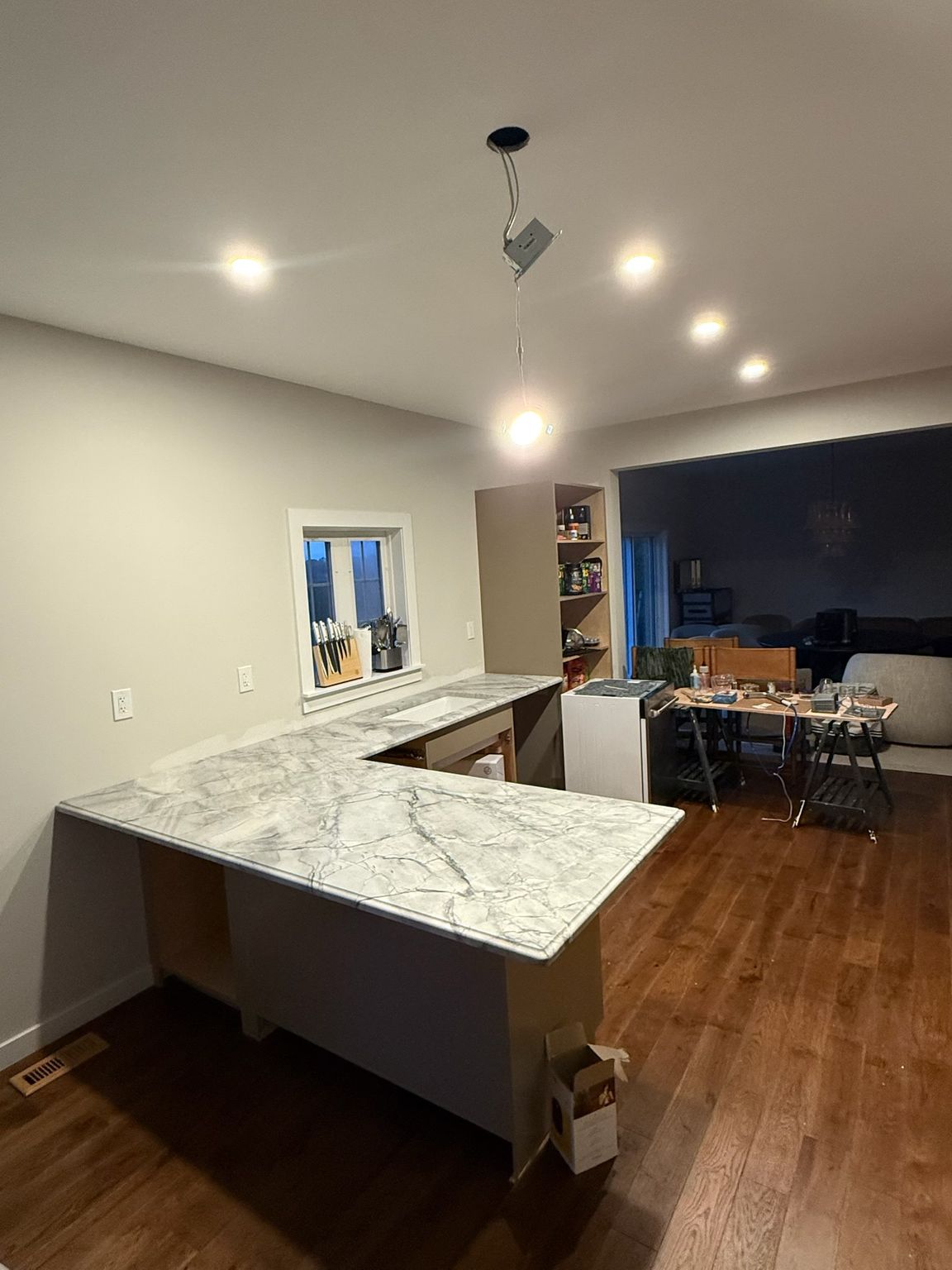 Modern kitchen with marble island, pendant light, and open view into a dark living room with TV and dining table