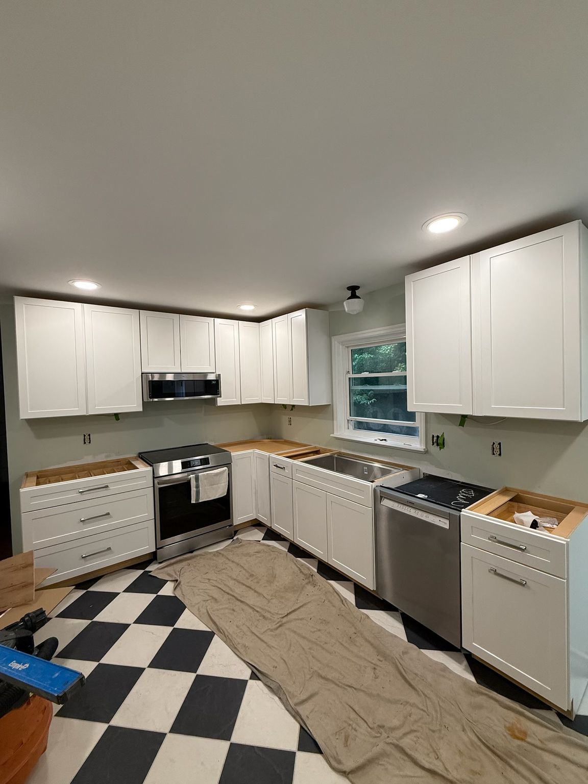 Small kitchen under renovation with white cabinets, black-and-white tile floor, and covered floor area.