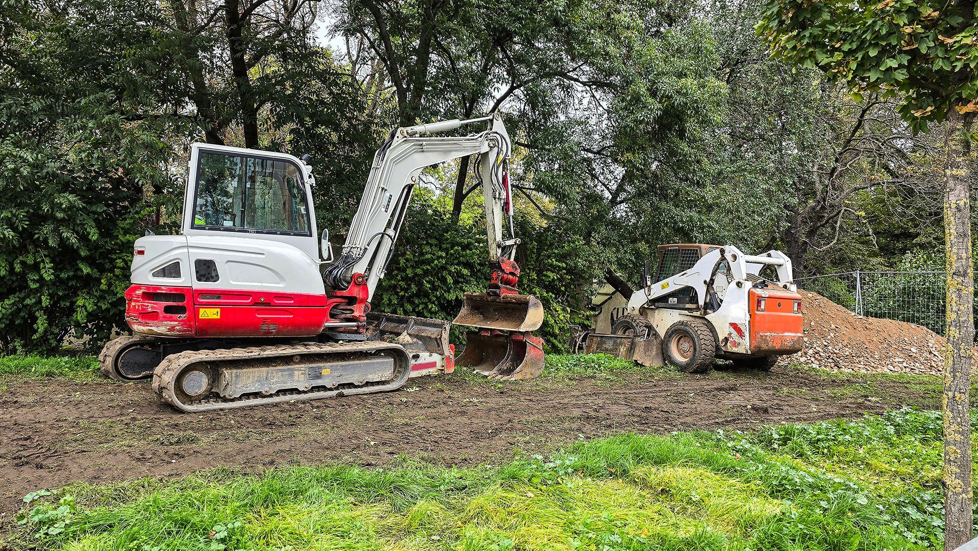 Excavator and skid steer on muddy ground, trees in background, likely construction site.