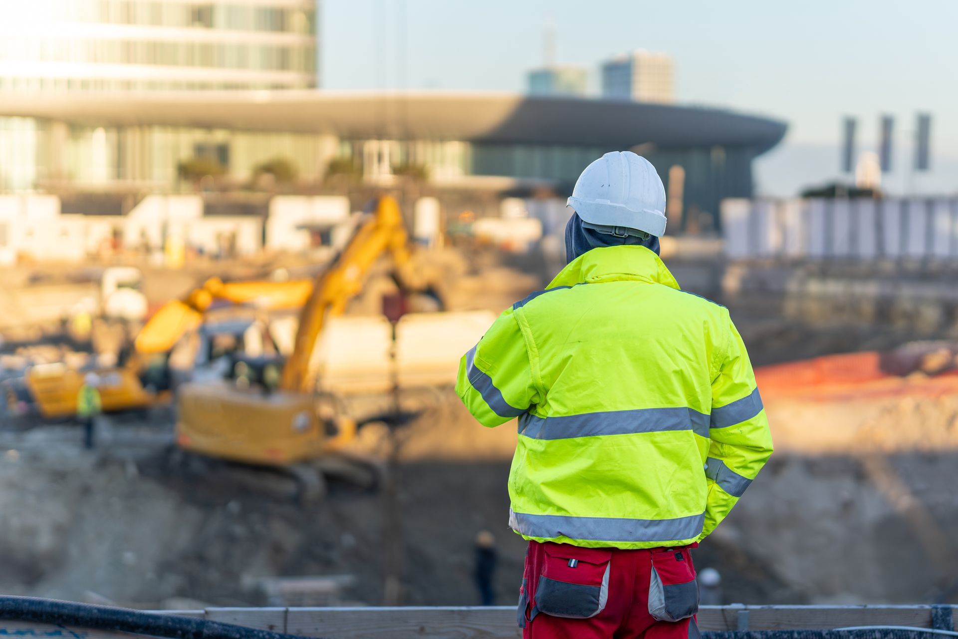 A construction worker in a high-visibility jacket and hard hat overseeing an excavator at a site. A construction worker in a high-visibility jacket and hard hat overseeing an excavator at a site.