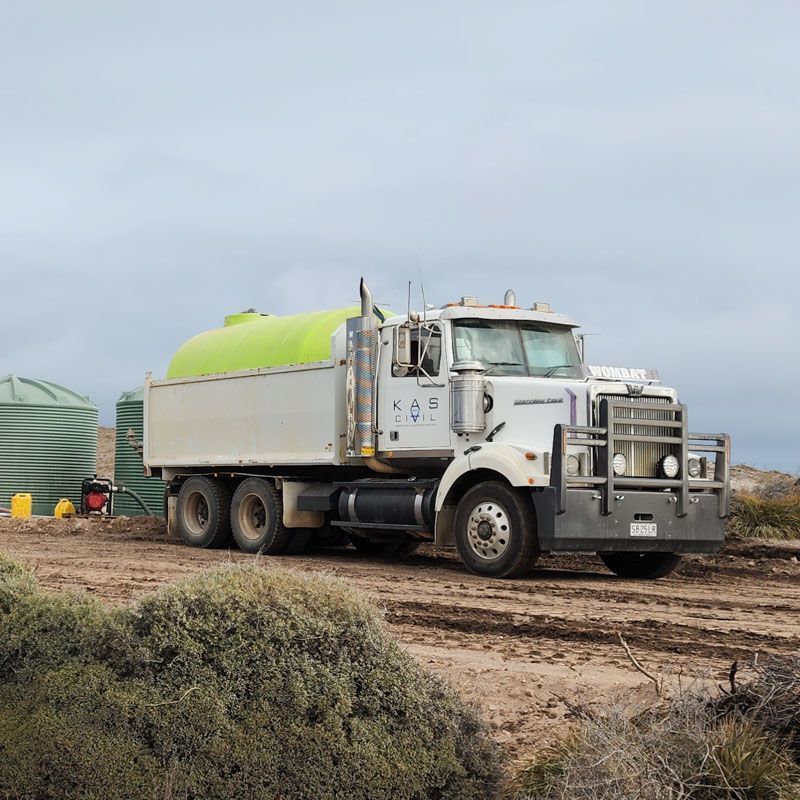 A white water tanker truck parked near green tanks on a dirt road, under a cloudy sky.