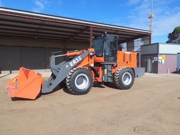 Orange ER32 wheel loader with raised bucket, parked on dirt near a building, against a blue sky.