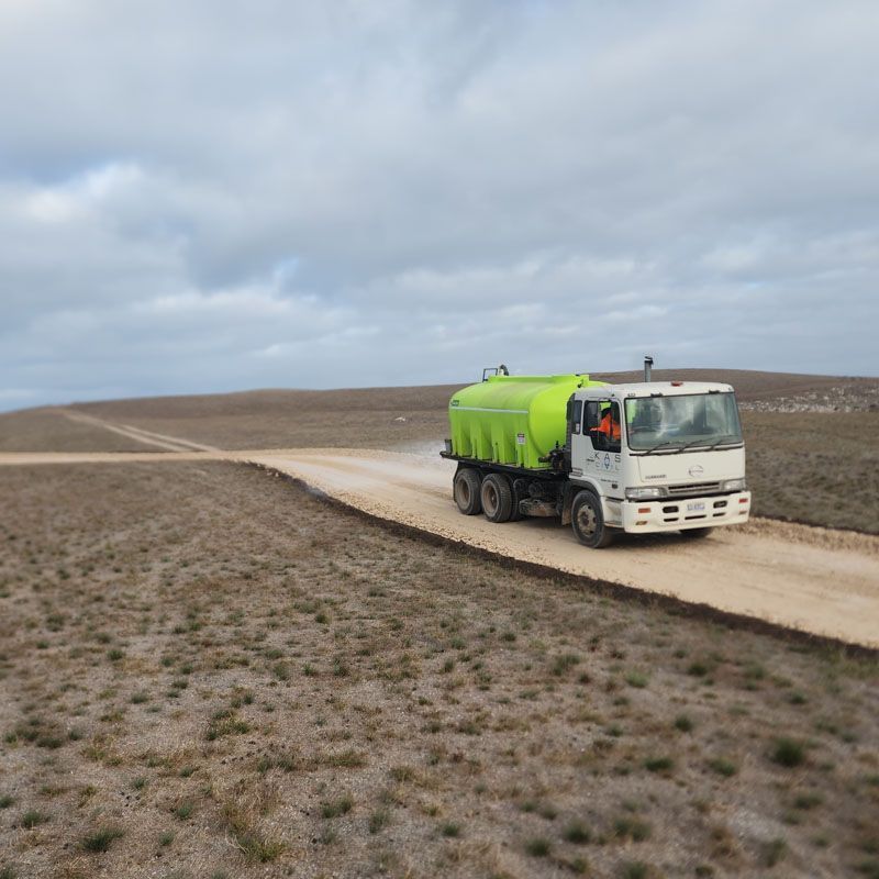 A water truck sprays a dirt road in a dry, open landscape under a cloudy sky.