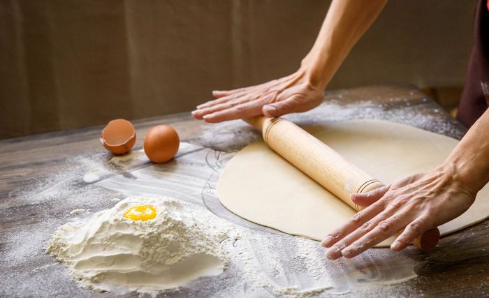 Mani che stendono l'impasto con un mattarello di legno su un piano di lavoro infarinato; uova e farina nelle vicinanze.
