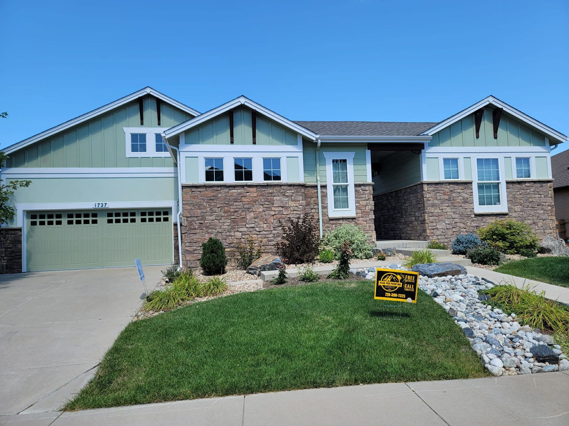 A single-story suburban house with sage green siding, stone veneer accents, a multi-car garage, and a manicured front lawn.