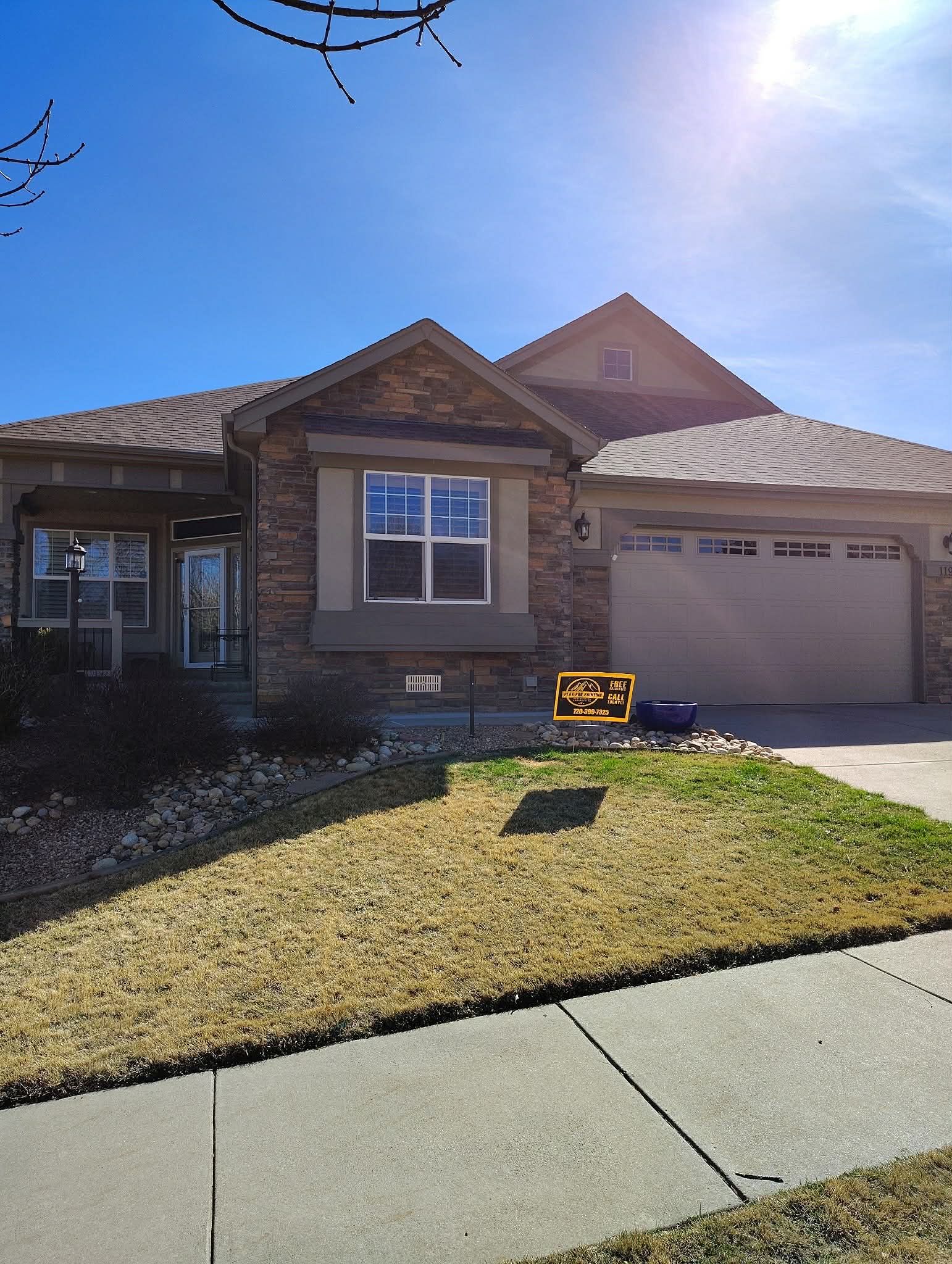 Single-story stone and beige house with a two-car garage and a small front lawn under a clear blue sky.