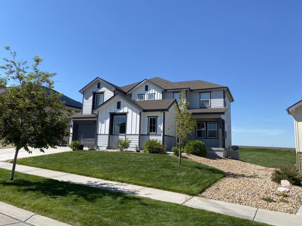 A two-story suburban house with a gray and white exterior, a green lawn, and a stone gravel side yard under a clear sky.