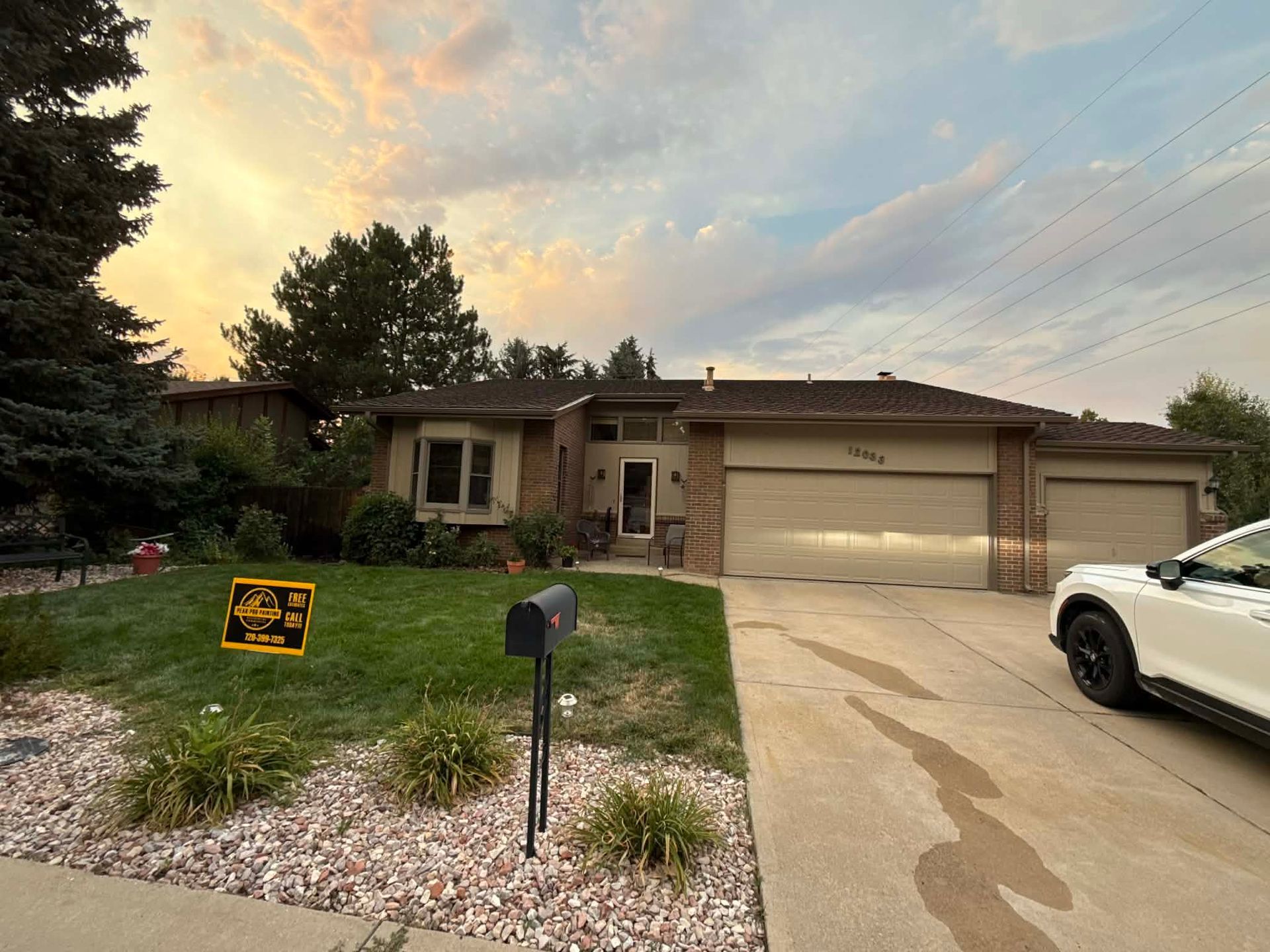 Single-story suburban house with a two-car garage, driveway, front lawn, and a parked white car at sunset.