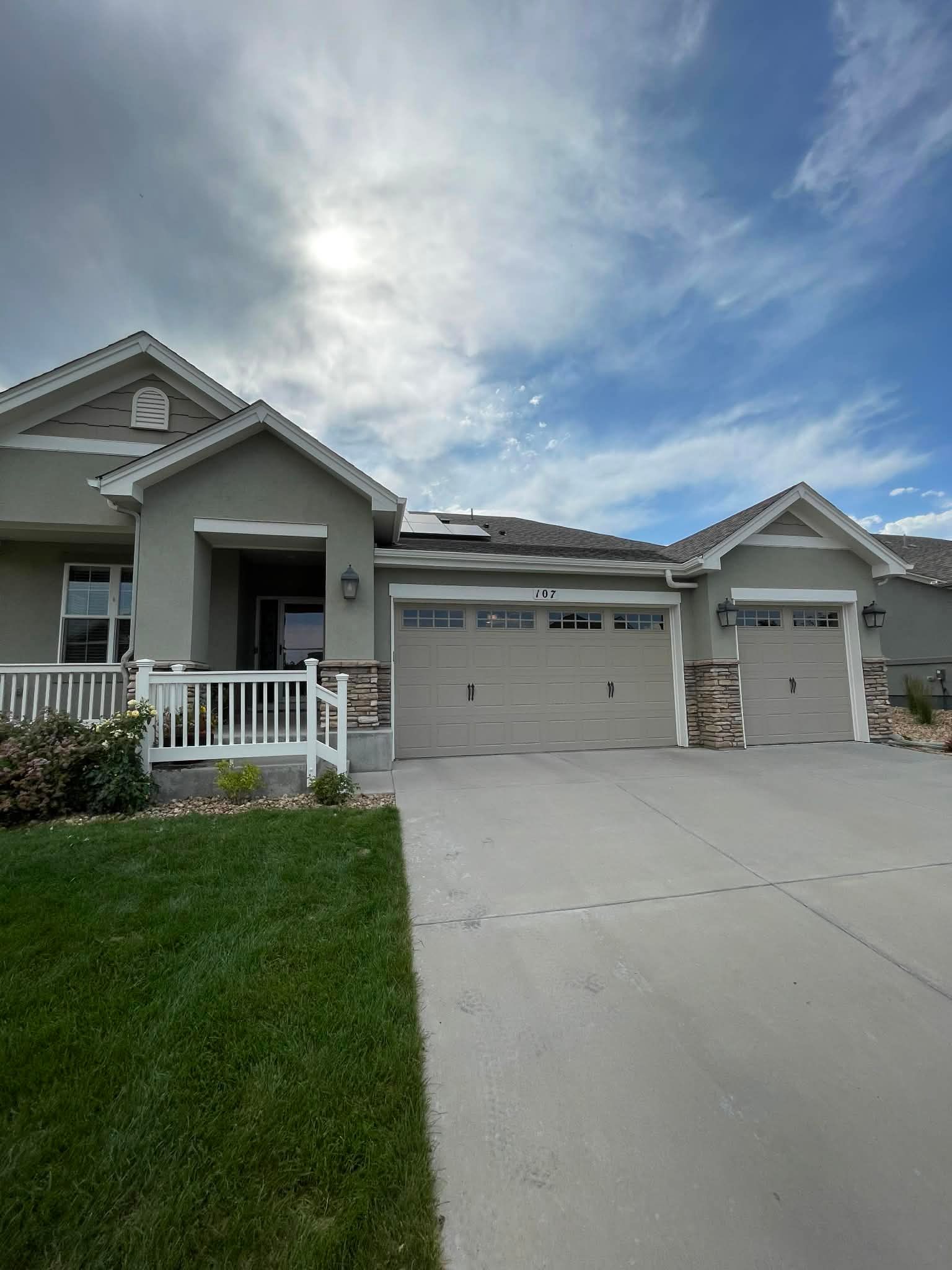 A modern one-story home with a tan exterior, stone accents, a three-car garage, and a white front porch railing.