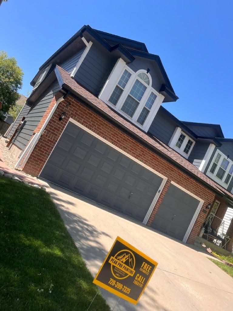A two-story house with grey siding and red brick accents, featuring a two-car garage and a small sign in the driveway.