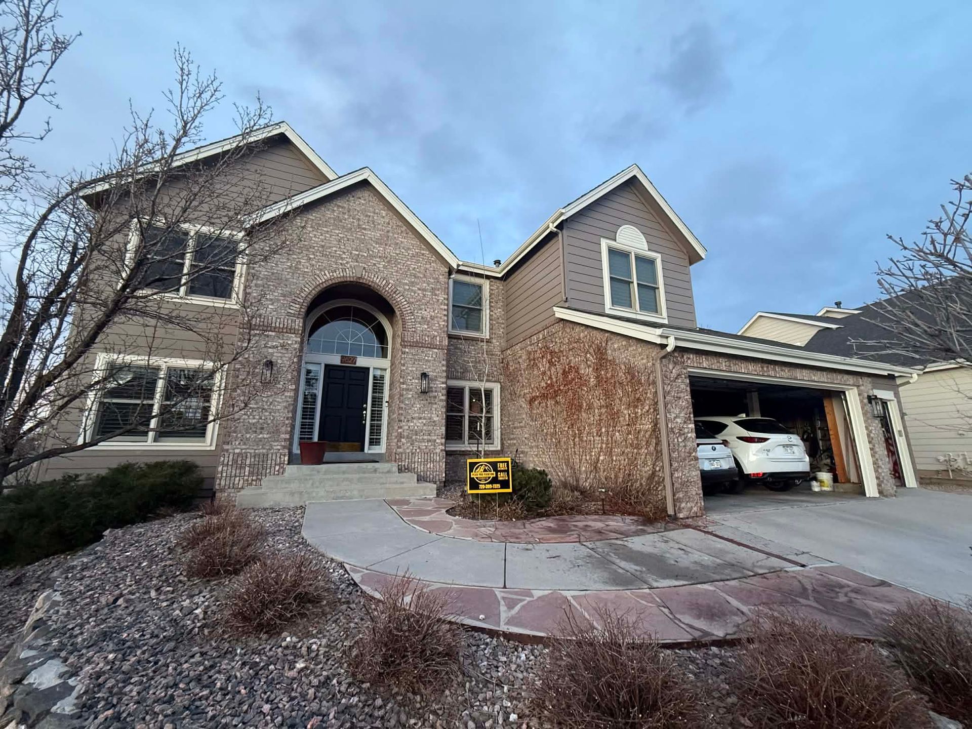 A multi-story brick suburban house with a gray shingle roof, arched entryway, and attached garage during twilight.
