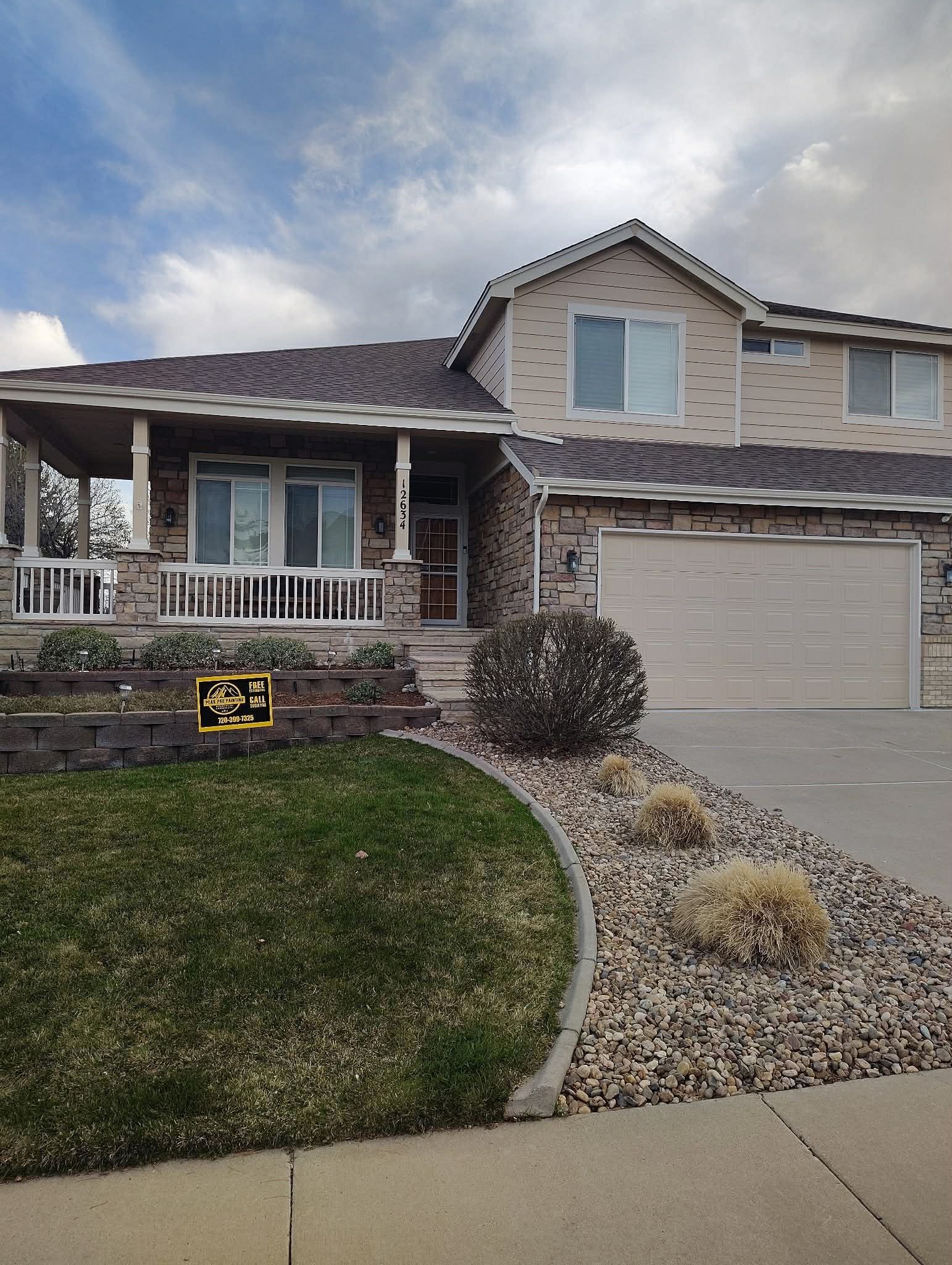 A two-story suburban home with tan brick, a dark shingled roof, a front porch, and a gravel yard with patches of grass.