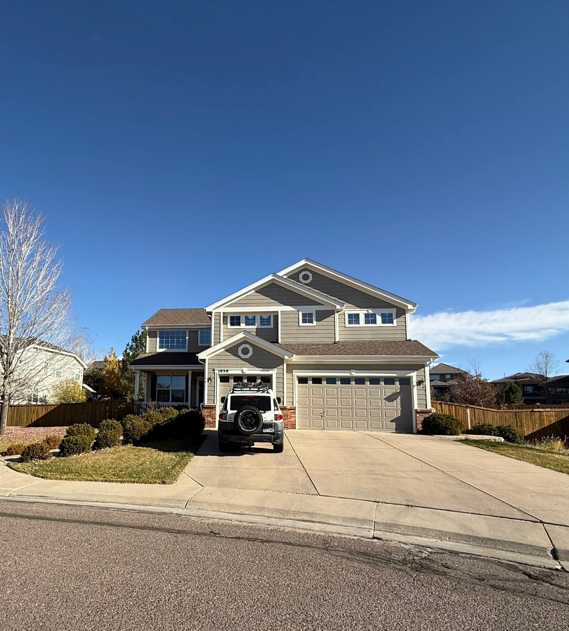 Two-story gray suburban house with a driveway, parked vehicle, and clear blue sky