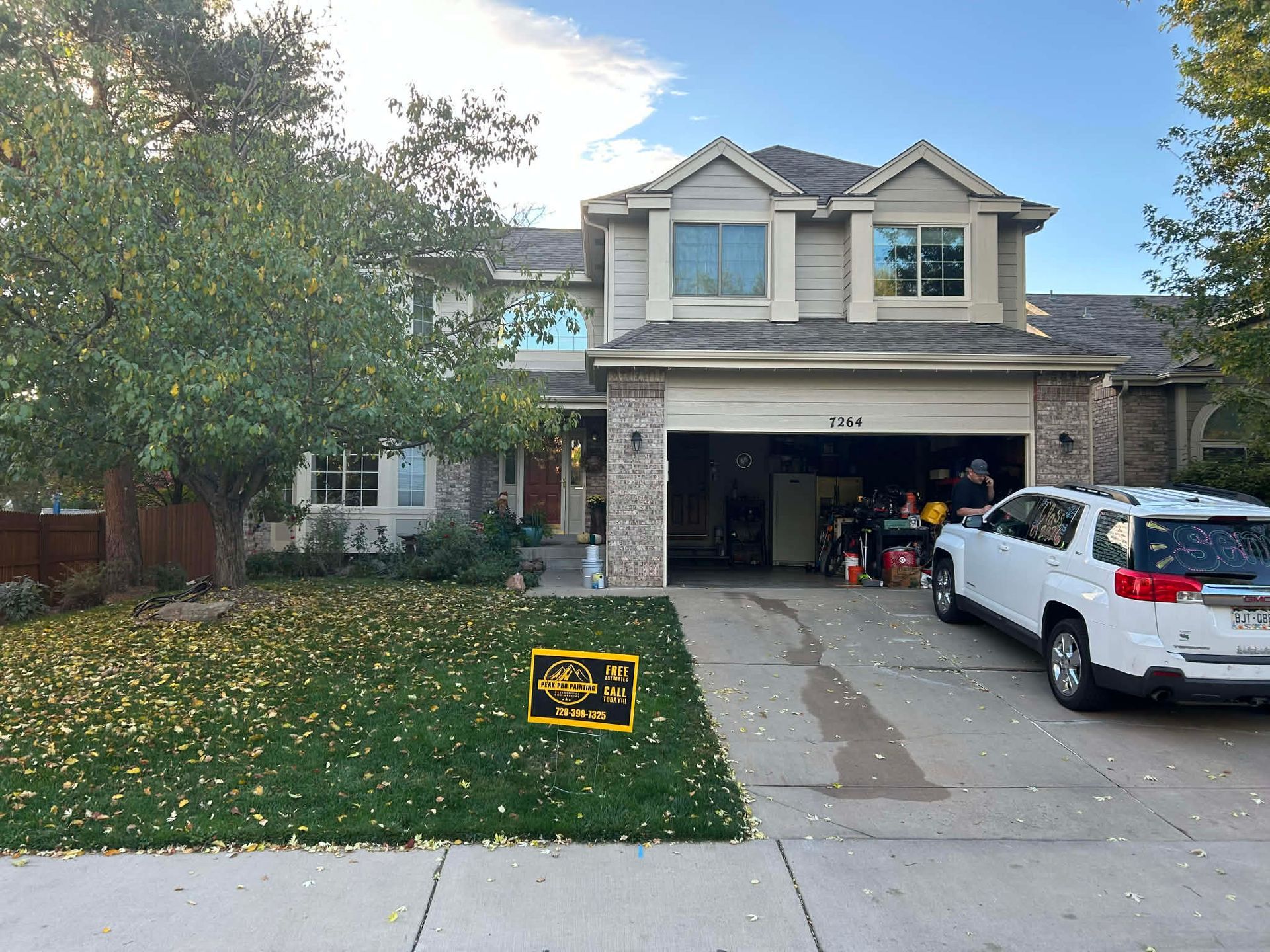 A two-story suburban home with tan siding and brick, featuring a lawn with fallen leaves and a white SUV in the driveway.