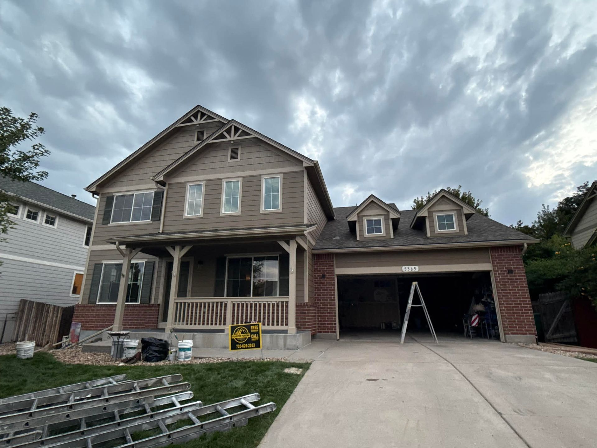 A tan two-story house with a brick base, covered front porch, and open garage under a cloudy sky with ladders on the lawn.