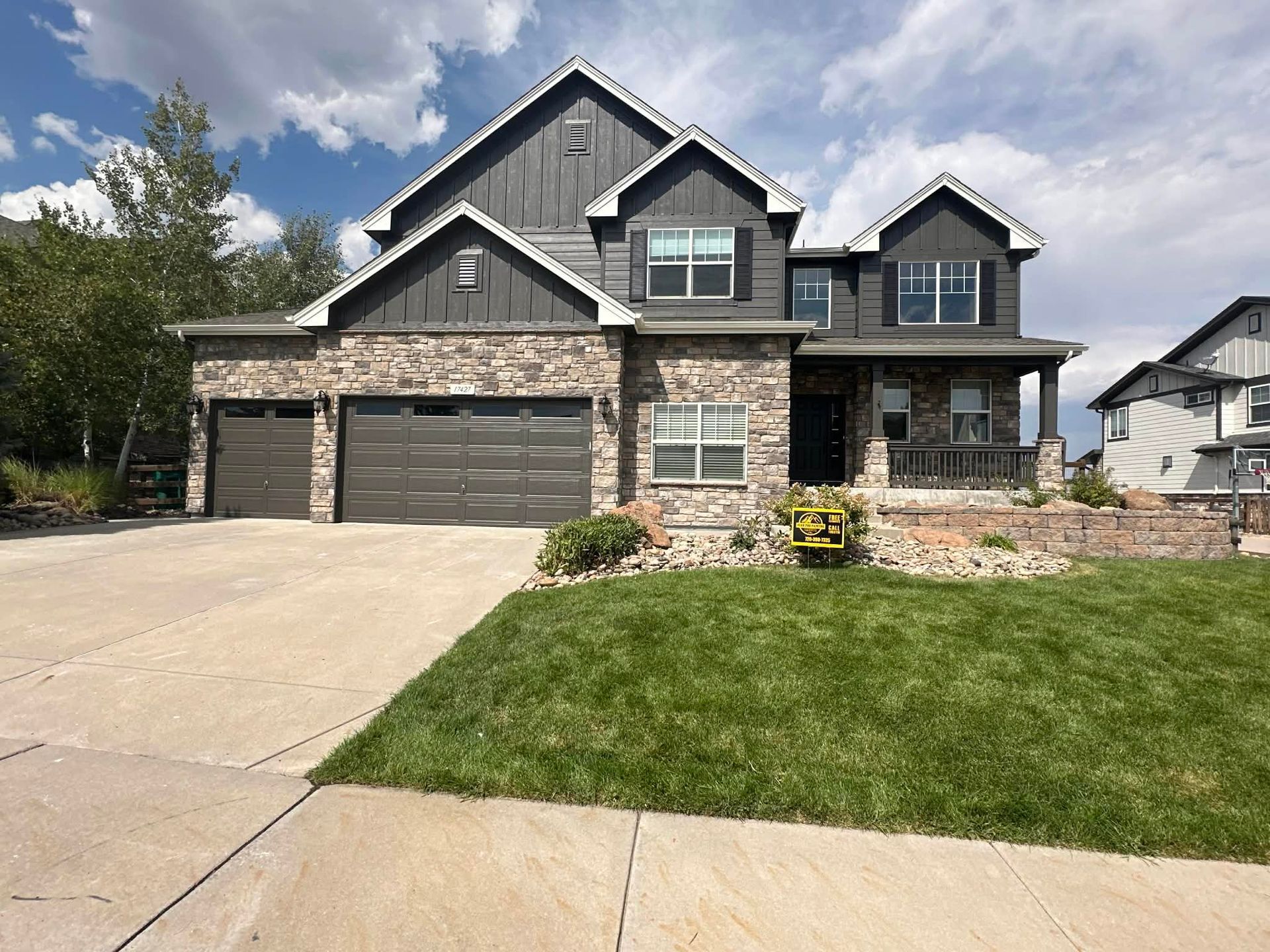 Two-story gray stone suburban house with a double garage, driveway, and front lawn under a cloudy sky