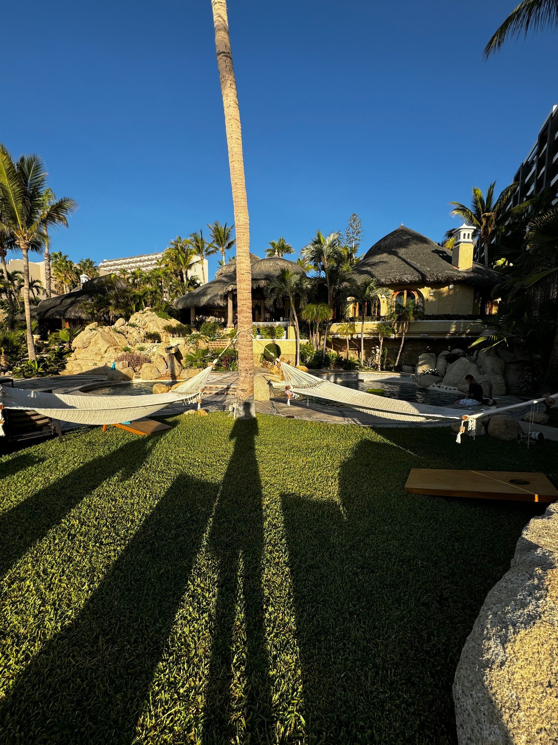 A lush green lawn with a palm tree, hammocks, and rustic huts under a clear blue sky, cast in long sunset shadows.