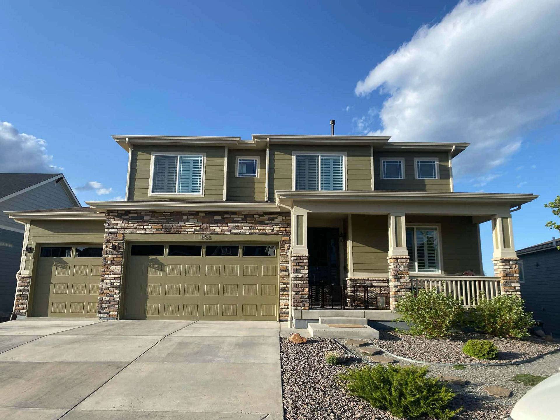 A two-story olive green suburban house with stone accents, a two-car garage, and a covered front porch under a blue sky.