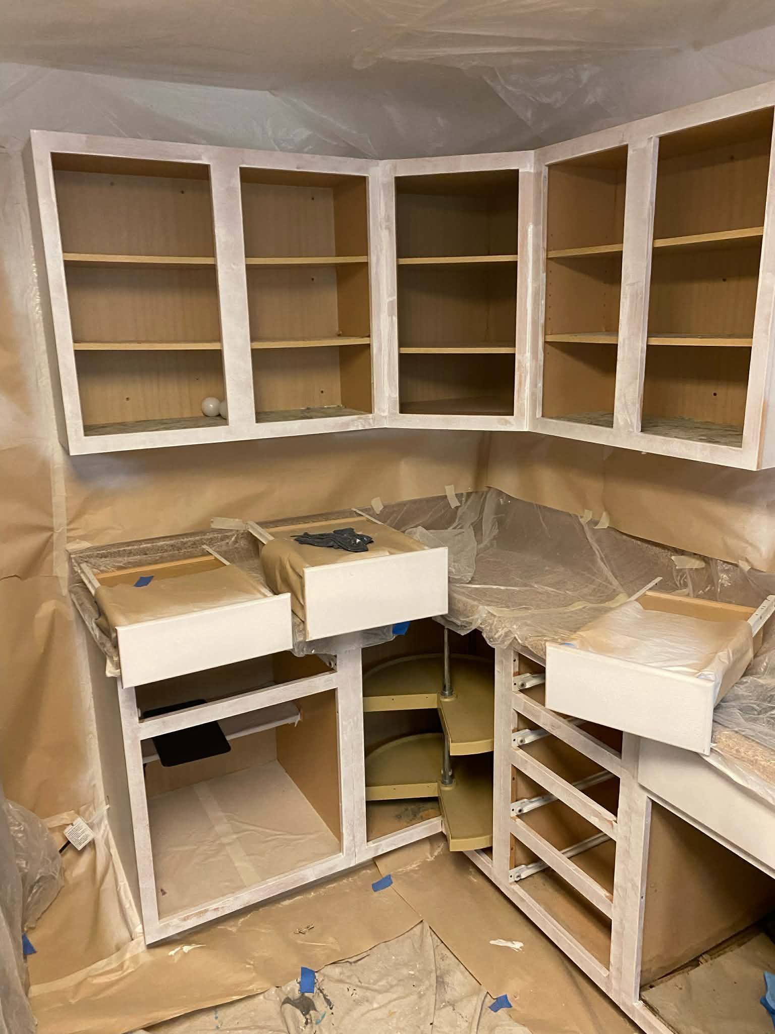 Kitchen cabinets being renovated, featuring open white-painted frames and paper-masked countertops in a construction zone.