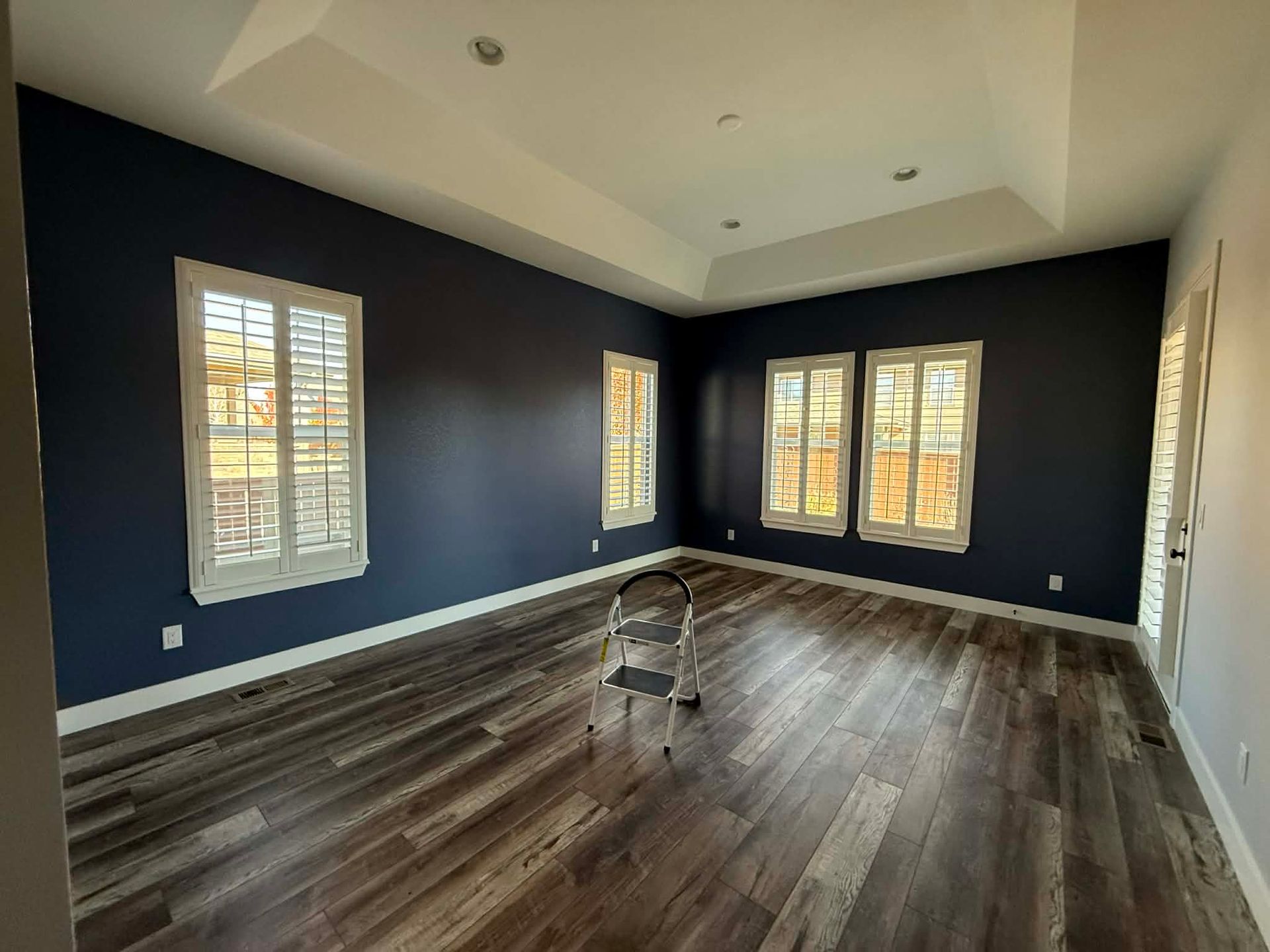 An empty room with dark blue walls, wood-look flooring, white trim, plantation shutters, and a small metal stepladder.