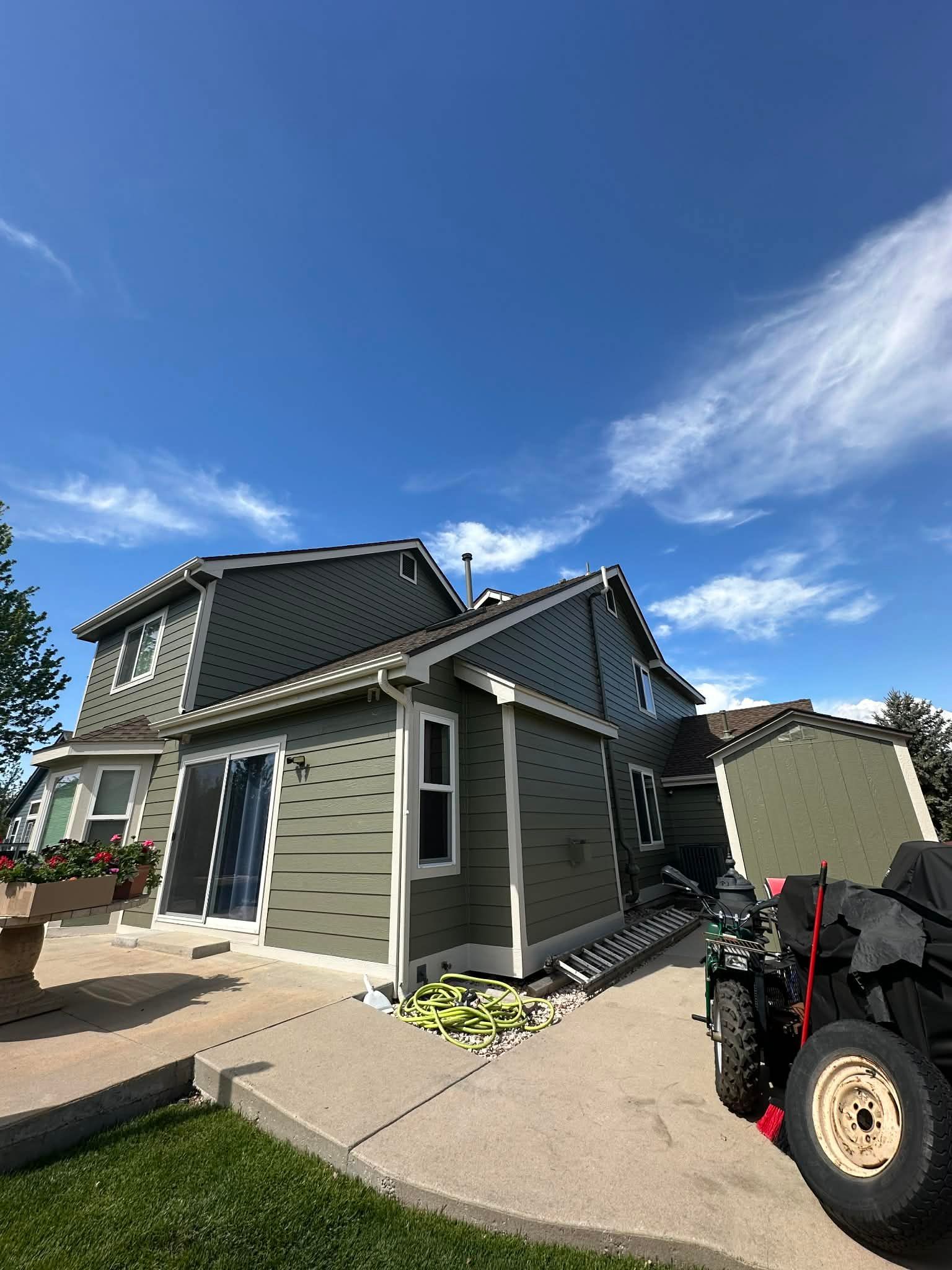 Gray house with dark roof under a blue sky, next to a trailer and tractor wheel on a paved path