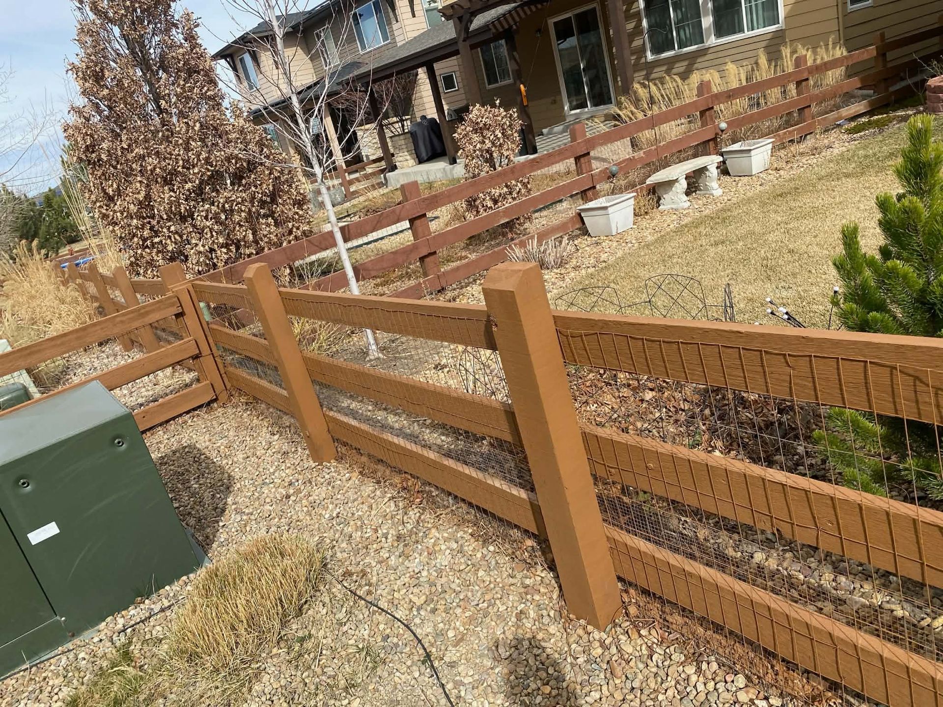 A wooden post-and-rail fence runs through a yard with gravel landscaping, near a utility box and a house.