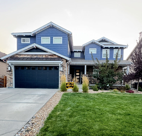 A two-story blue house with a dark garage, stone siding, and a neatly manicured front lawn under a bright sky.
