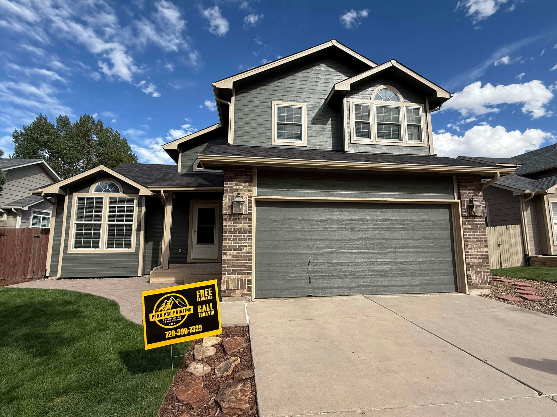 A two-story grey house with a stone-accented front, a large garage, and a lawn sign under a blue, cloudy sky.