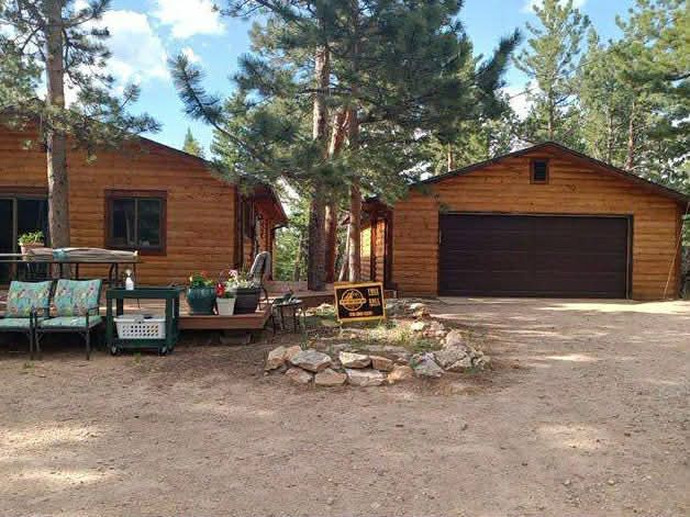 Wooden cabin and detached garage in a pine forest clearing with a gravel drive and patio seating.
