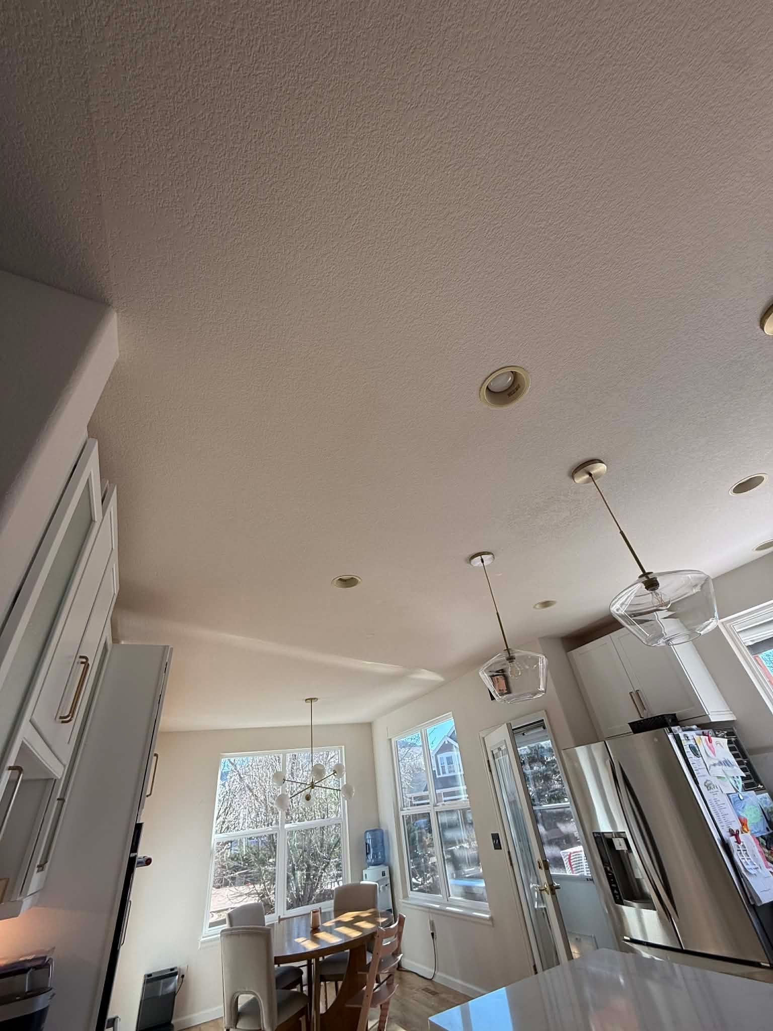 A dining area featuring a table, chairs, windows with natural light, recessed ceiling lights, and two pendant fixtures.