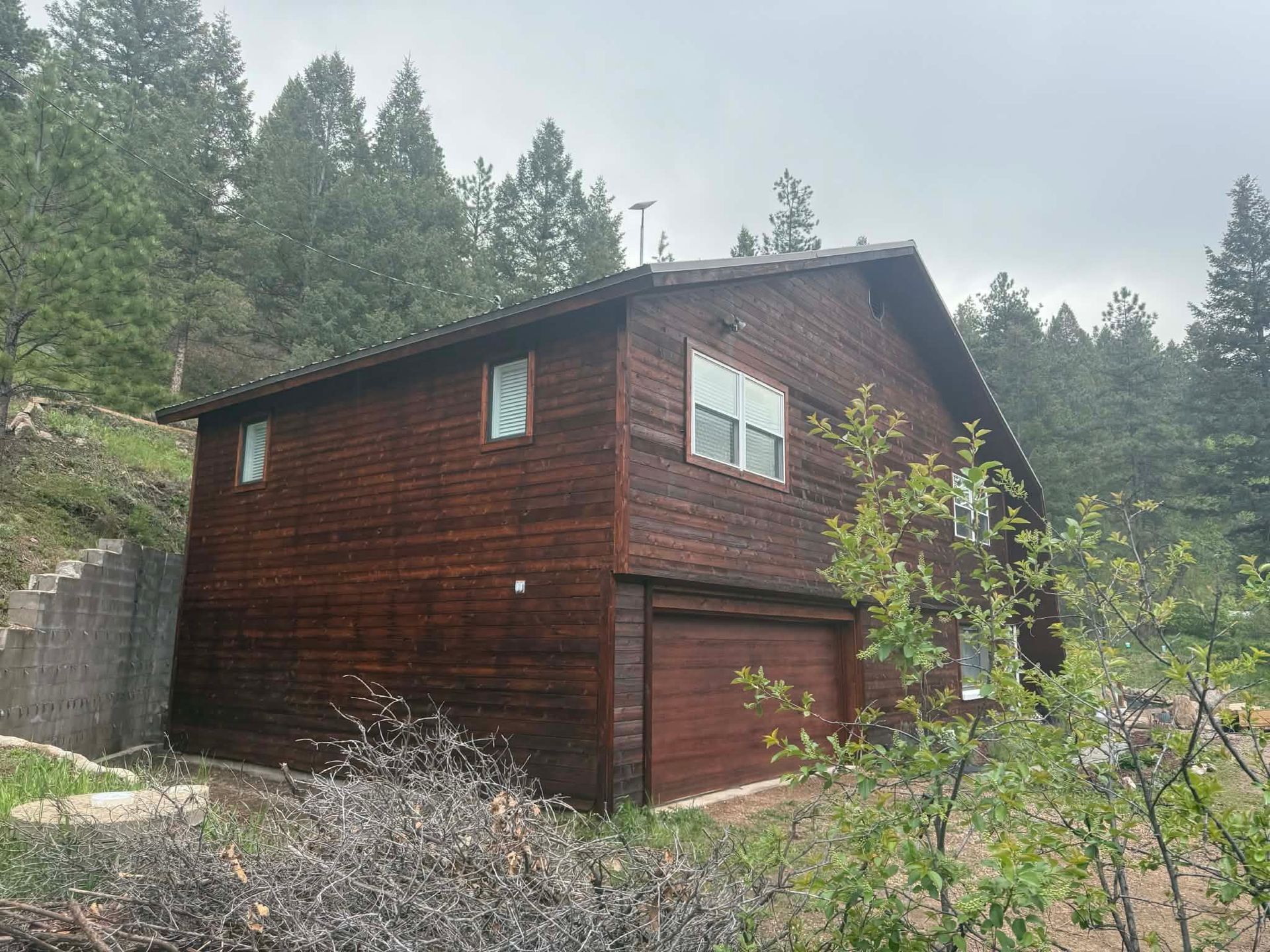 Dark wooden cabin with garage door in a forested, rocky hillside setting