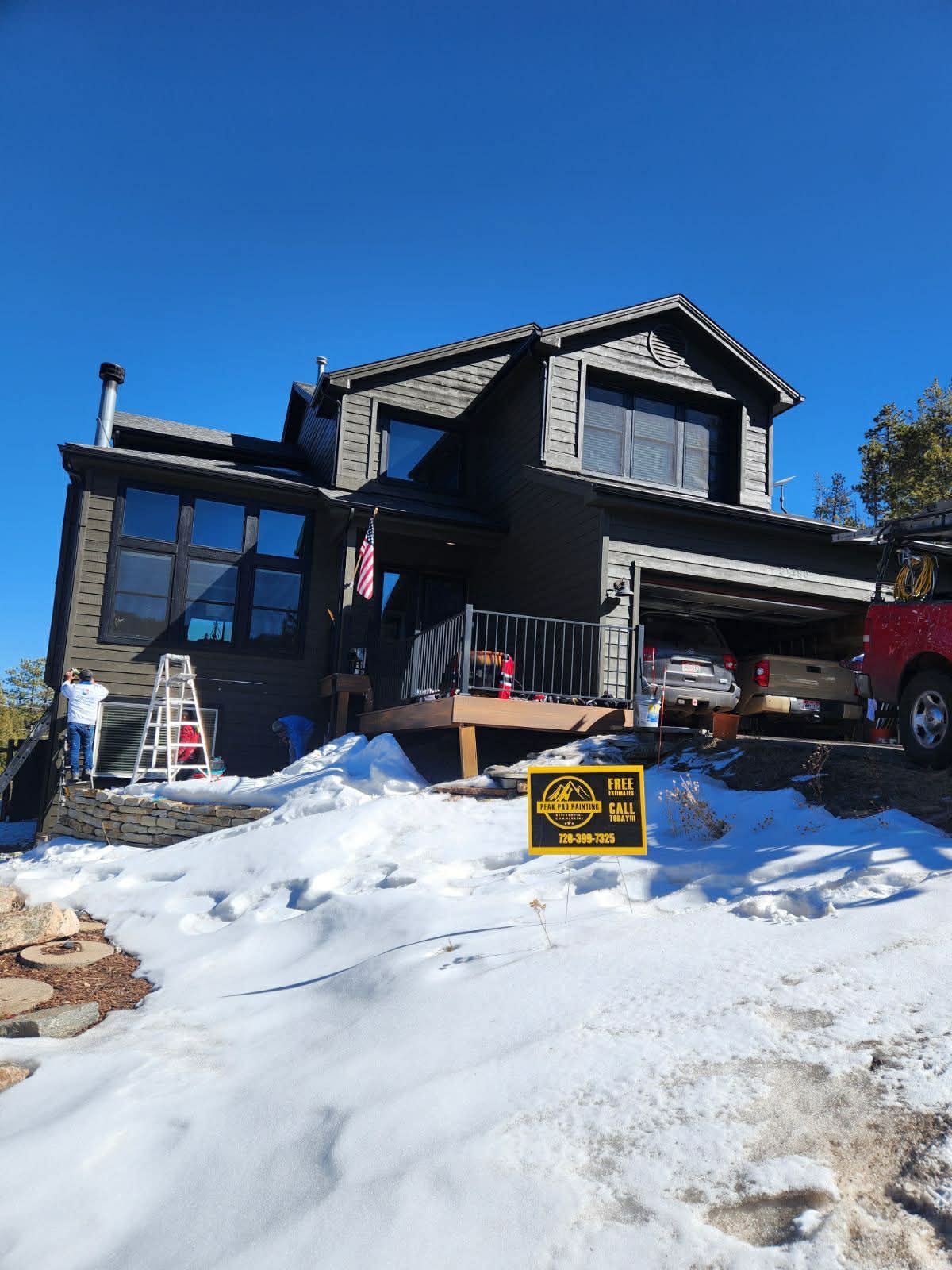 A dark two-story house with a garage and a ladder on a snowy, sunny day, with a construction sign in the foreground.
