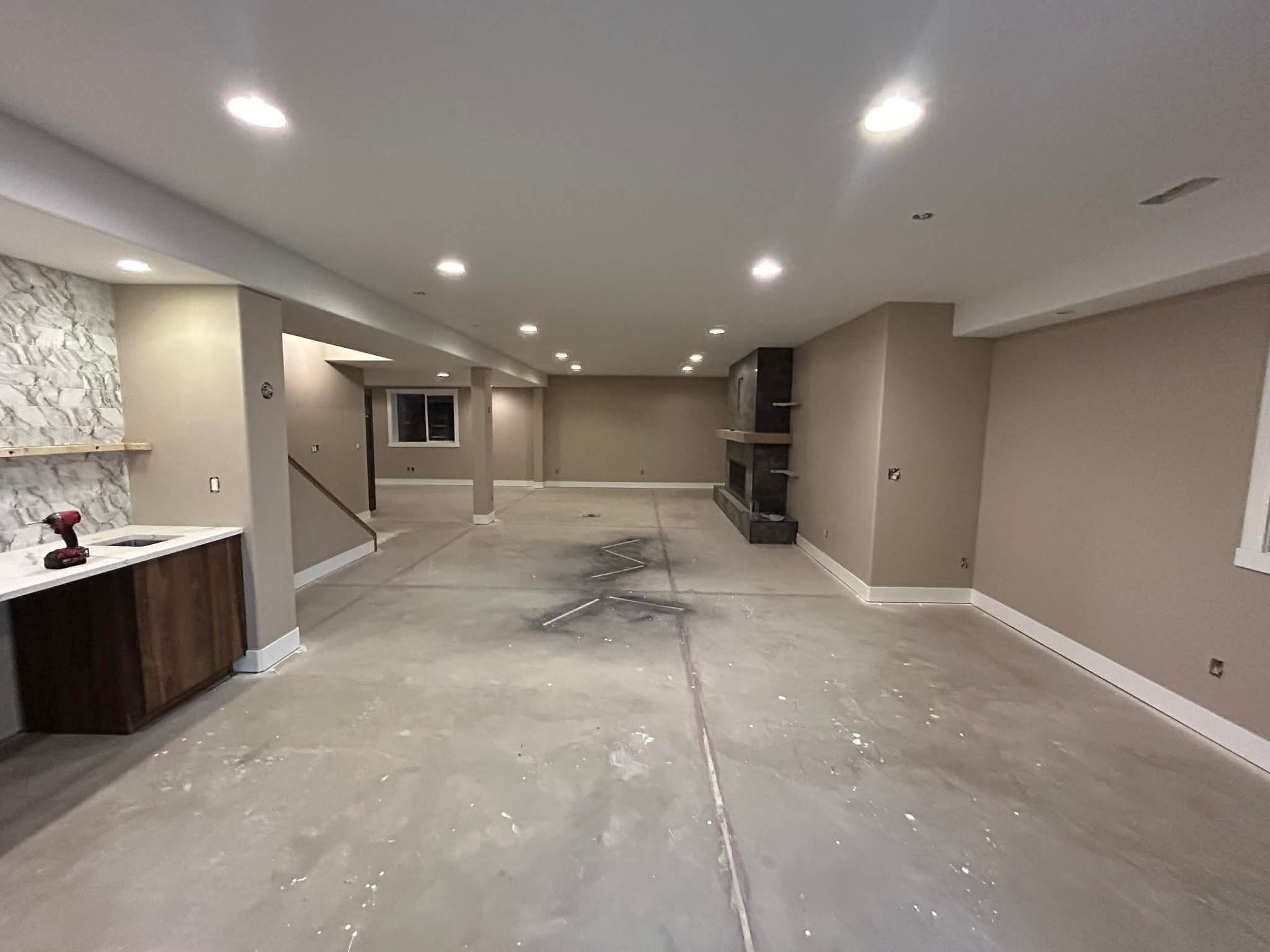 A spacious, unfinished basement with taupe walls, recessed lighting, a fireplace, and a small kitchenette area.