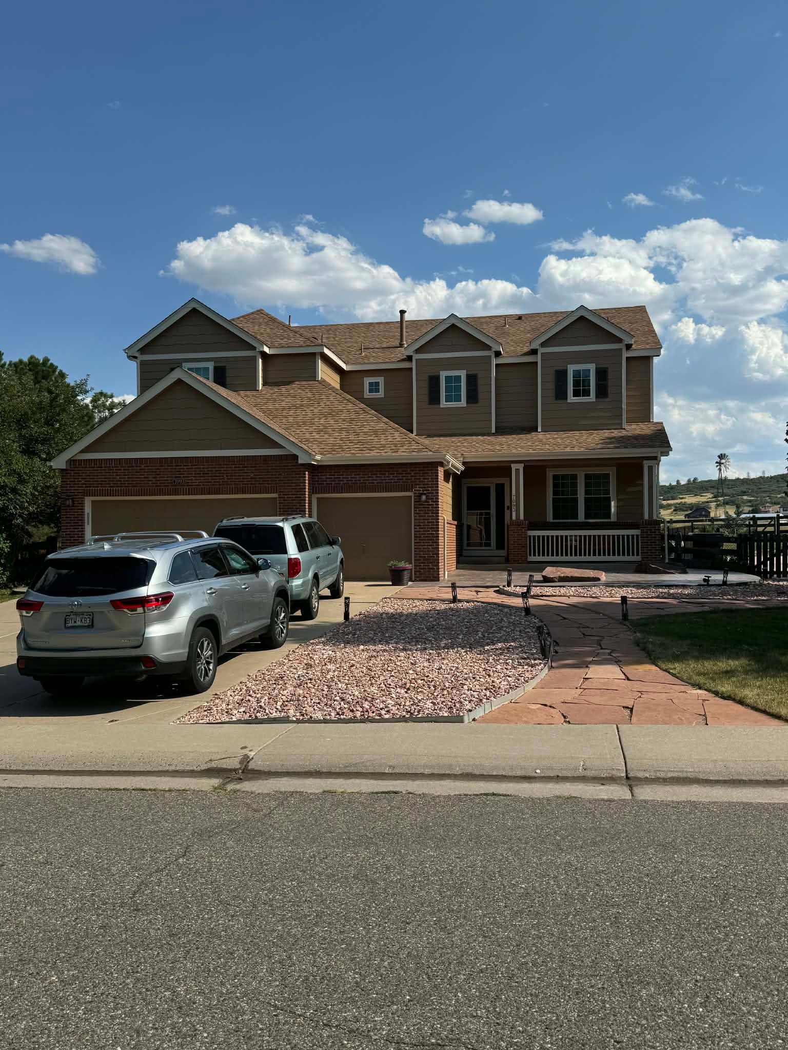 A two-story tan suburban house with a brown roof, brick accents, a two-car garage, and a light-colored stone driveway.