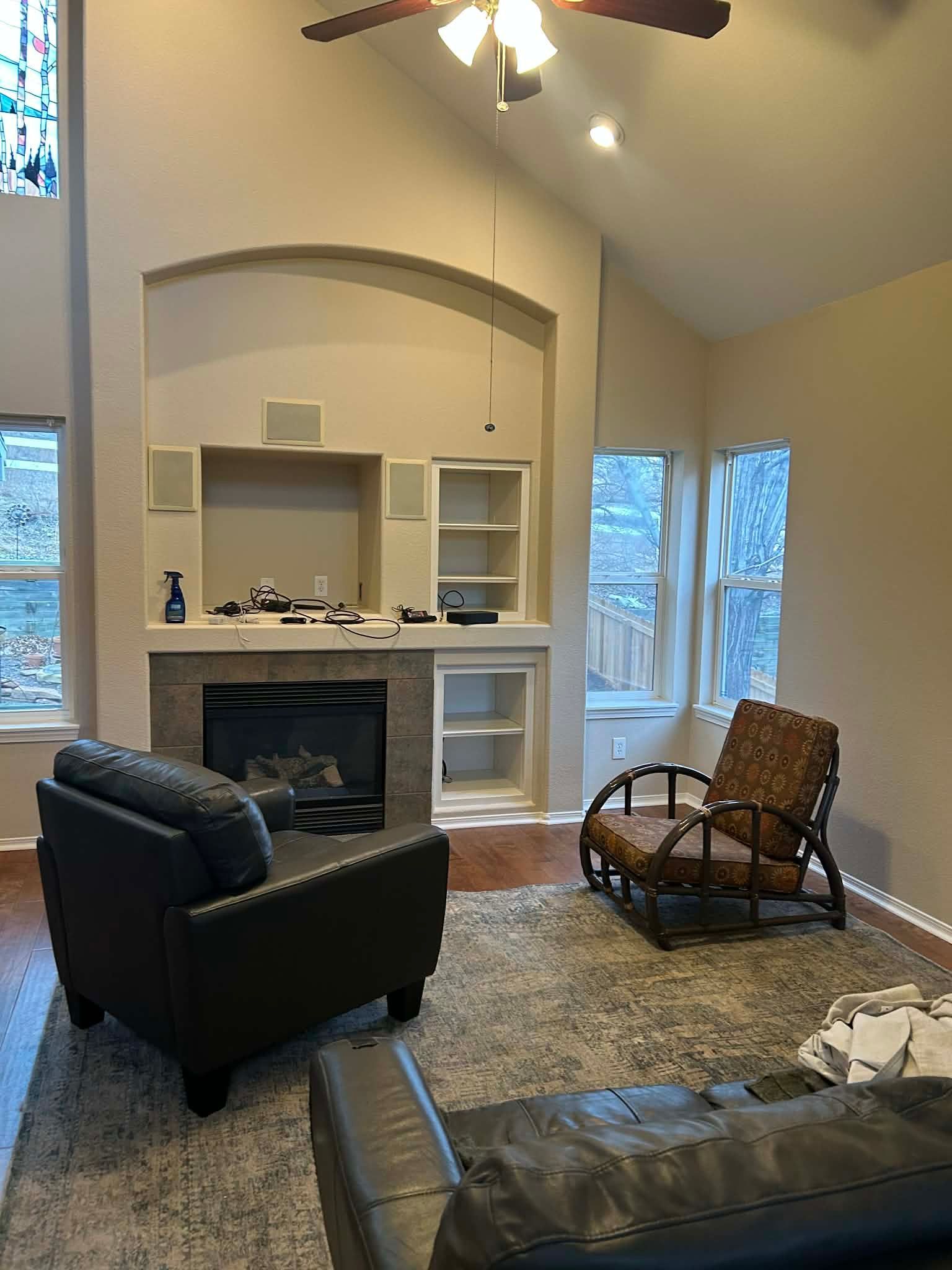 A living room with a stone fireplace, built-in shelving, two chairs, and a rug under a vaulted ceiling with a fan.