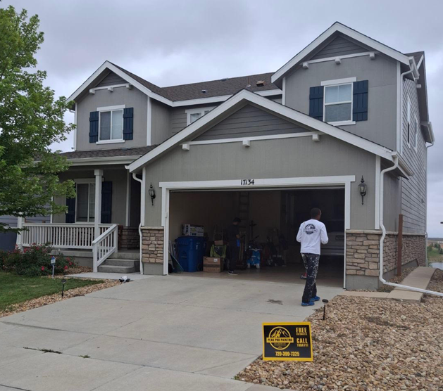 A two-story gray house with a garage and porch, featuring a person walking into the garage and a sign on the lawn.