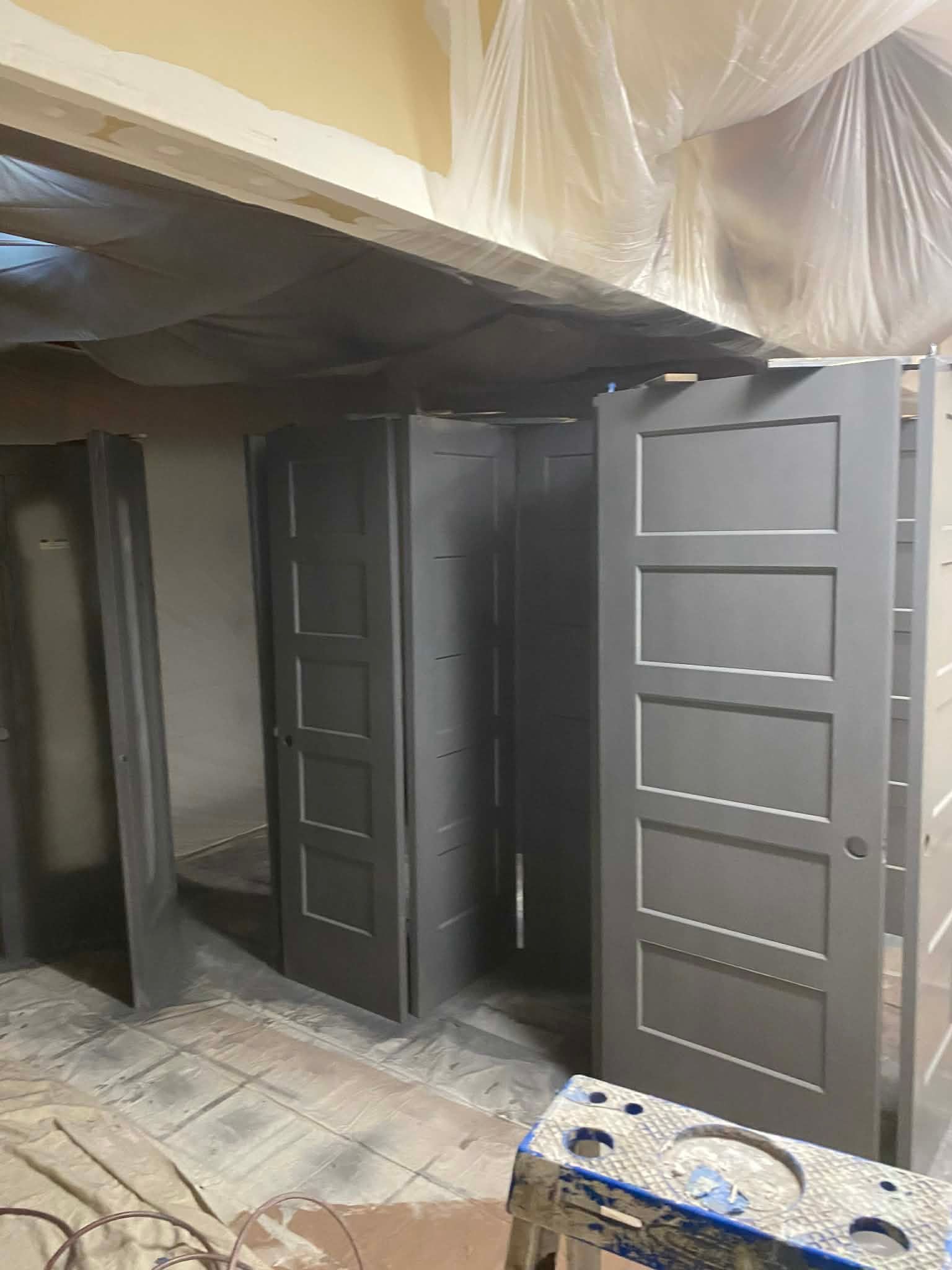 Several gray, five-panel doors standing in a row in a room under construction with plastic sheeting overhead.