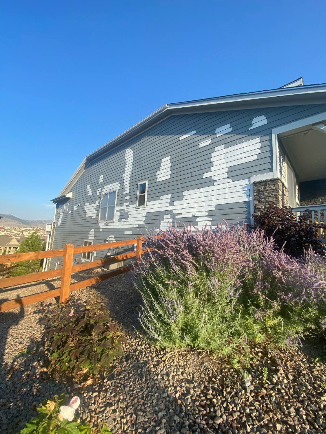 A side view of a house with light blue siding showing significant, patchy paint peeling against a clear blue sky.