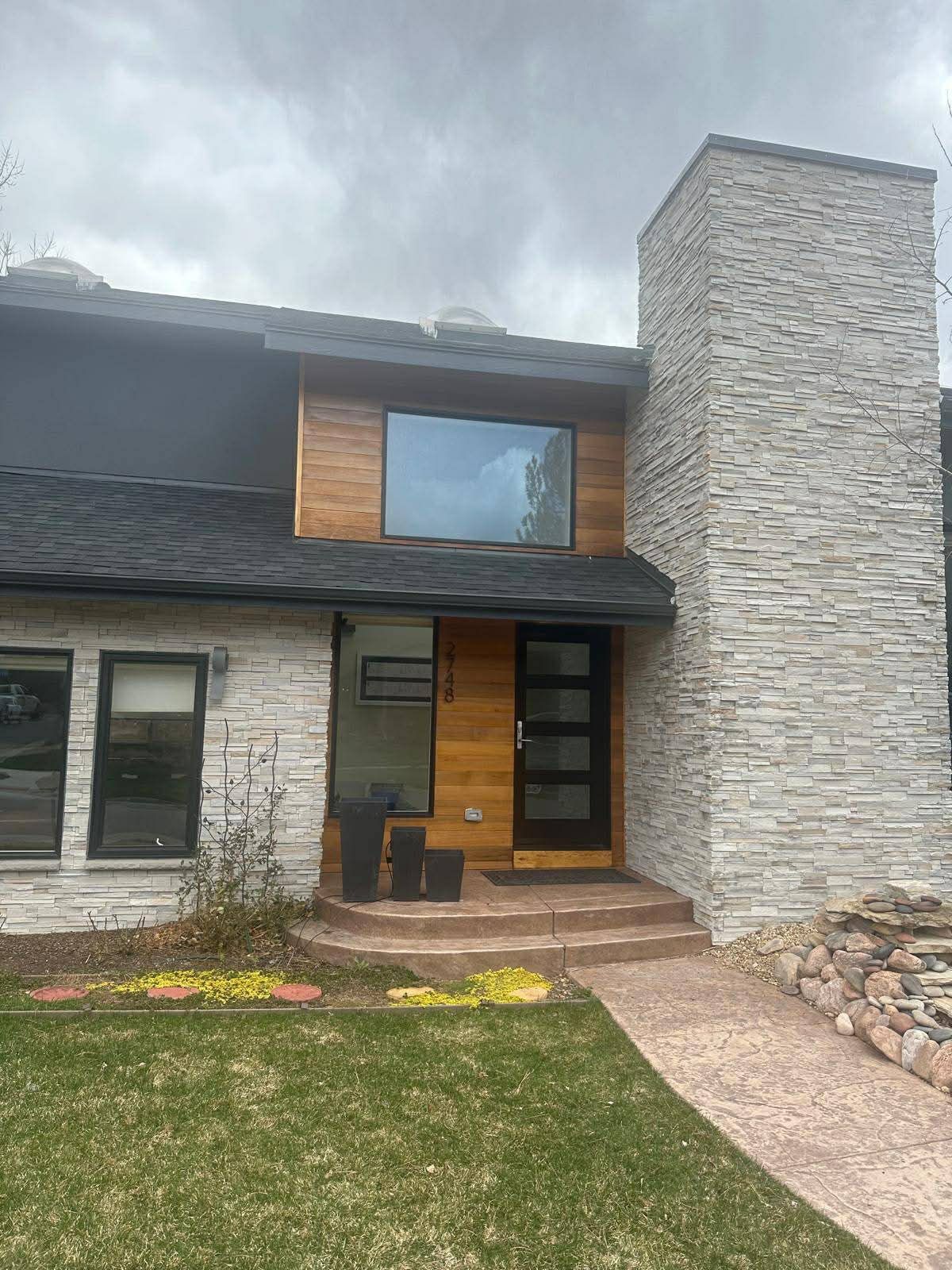 Modern stone-and-wood house exterior with front entry, chimney, lawn, and walkway